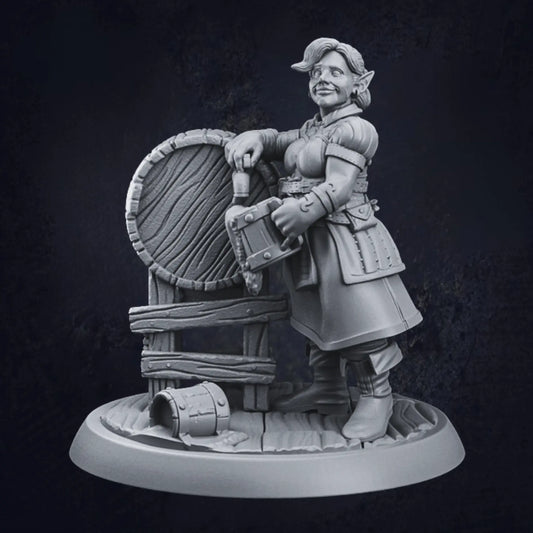 A miniature figurine of a female elven tavern keeper standing beside a large barrel. The figure wears medieval-style attire, including a belted tunic and boots, and holds a tankard in one hand. The other hand rests casually on the barrel, with a small bucket on the ground nearby. The figure is mounted on a textured, circular base, adding detail to the scene.