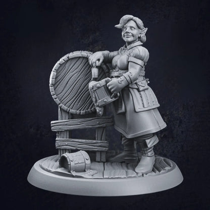 A miniature figurine of a female elven tavern keeper standing beside a large barrel. The figure wears medieval-style attire, including a belted tunic and boots, and holds a tankard in one hand. The other hand rests casually on the barrel, with a small bucket on the ground nearby. The figure is mounted on a textured, circular base, adding detail to the scene.
