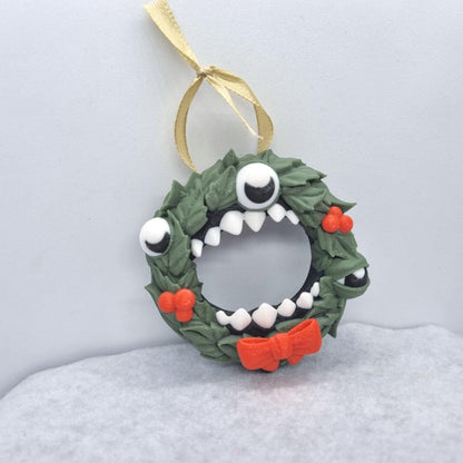 A whimsical wreath ornament with a monster-like face, featuring green leaves, red berries, and a red bow tie.